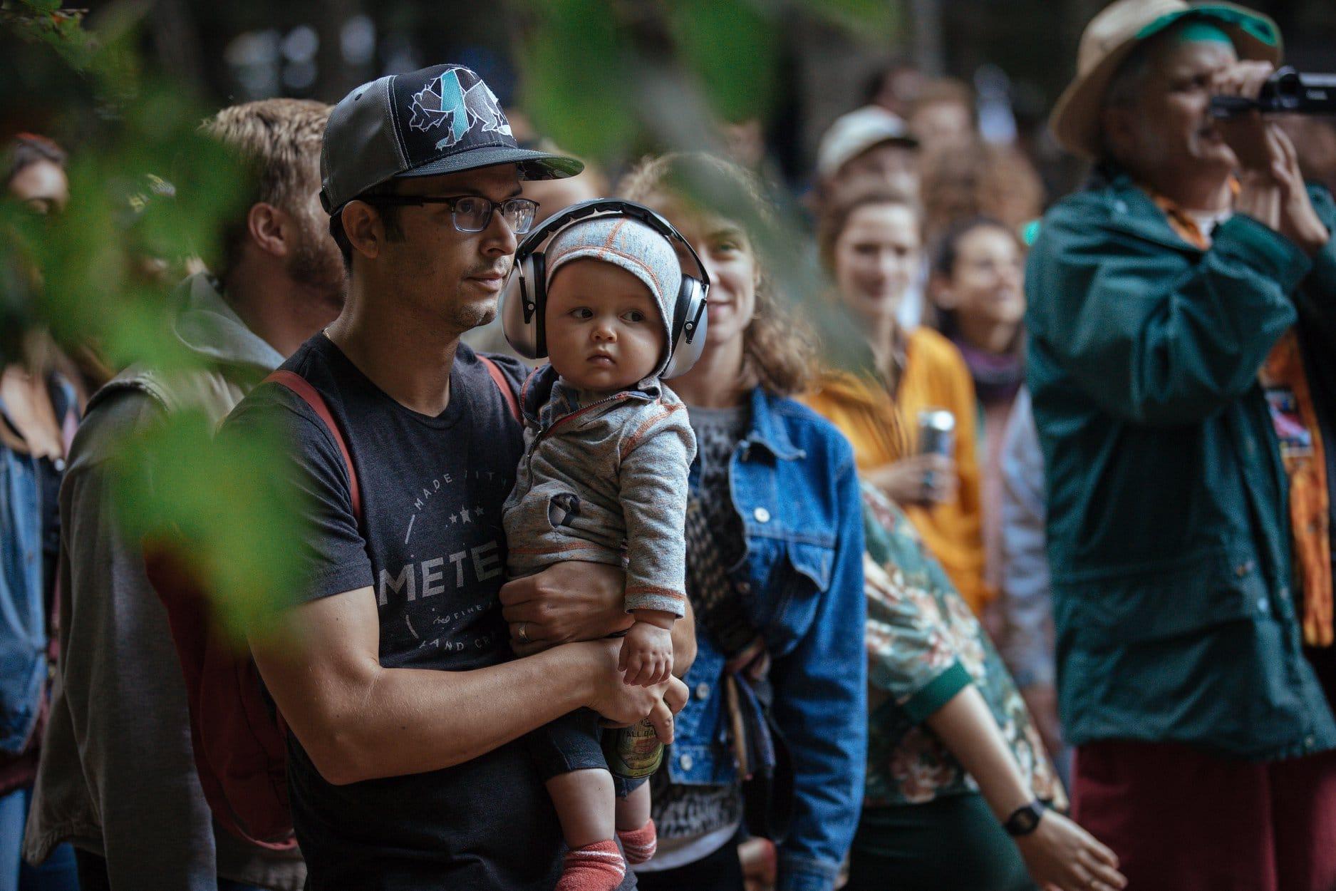 Joe holds his baby son, who wears ear protection, in a crowd watching an outdoor concert.