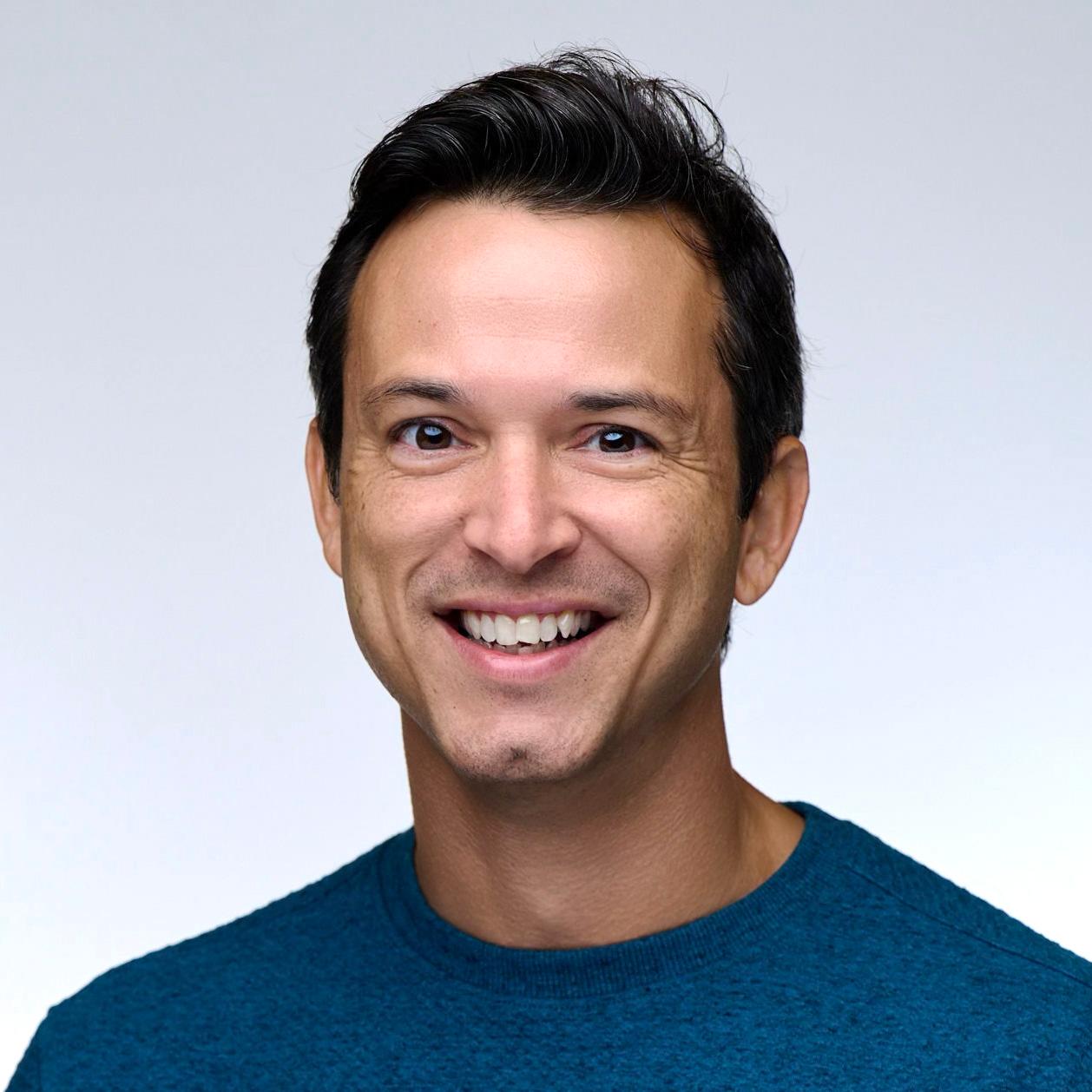 Headshot of Joe di Stefano smiling, wearing a teal sweater against a light gray background.