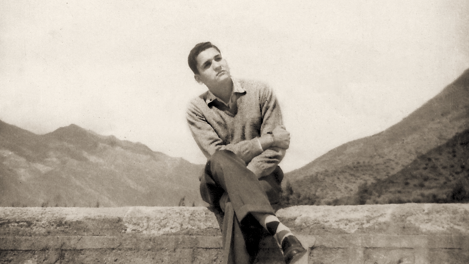 Sepia photograph of Javier Heraud as a young man in a sweater, sitting on a stone wall with mountains behind him.